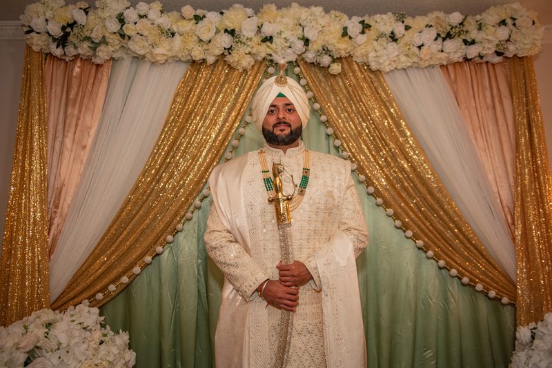 A formally dressed groom in white turban and embroidered sherwani holds a ceremonial sword against gold and white drapes.