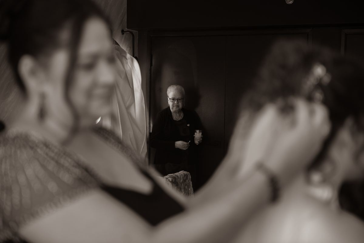 A woman in a doorway holds a cup while observing a bride and bridesmaid in preparation, capturing anticipation.