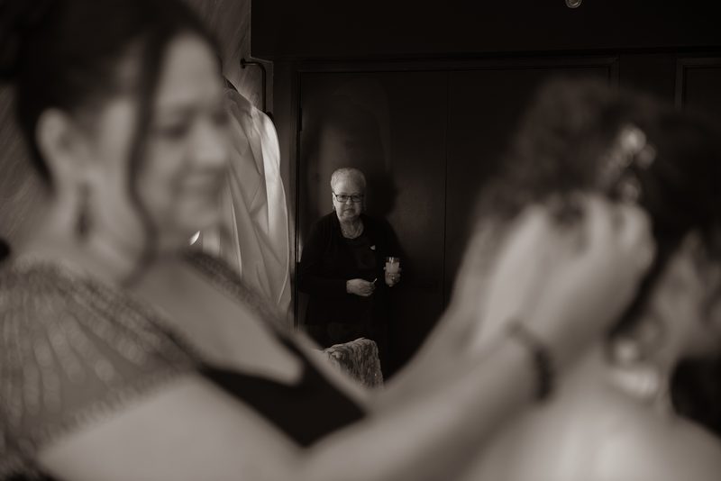 A woman in a doorway holds a cup while observing a bride and bridesmaid in preparation, capturing anticipation.