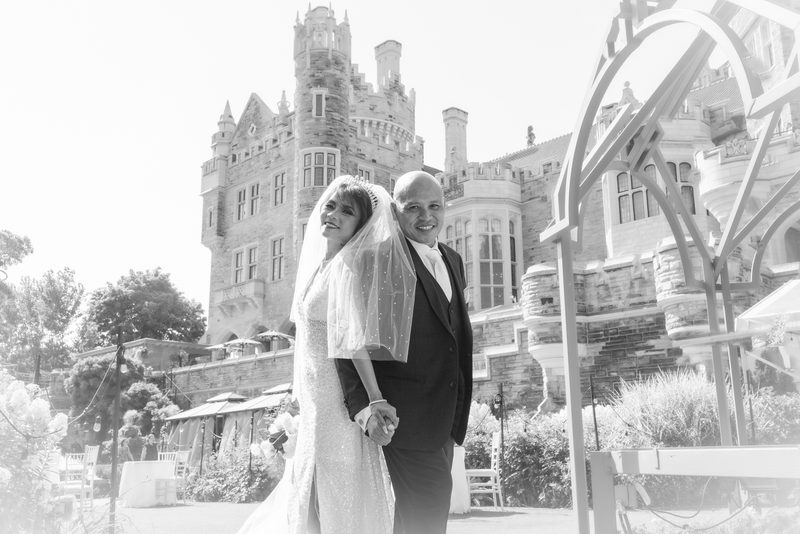 A bride and groom stand smiling in front of a stone building with an ornate metal arch partially framing them.