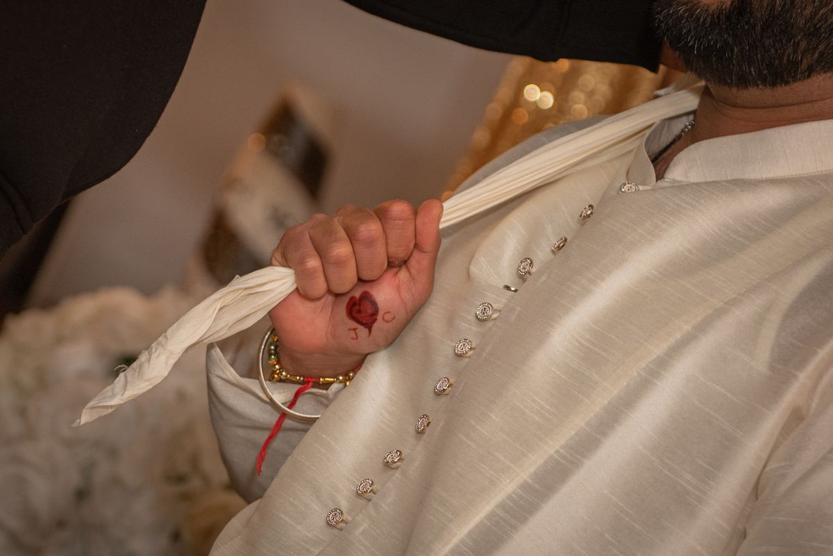A groom's hand holds a white cloth with a red heart and initials, wearing traditional attire in warm lighting.