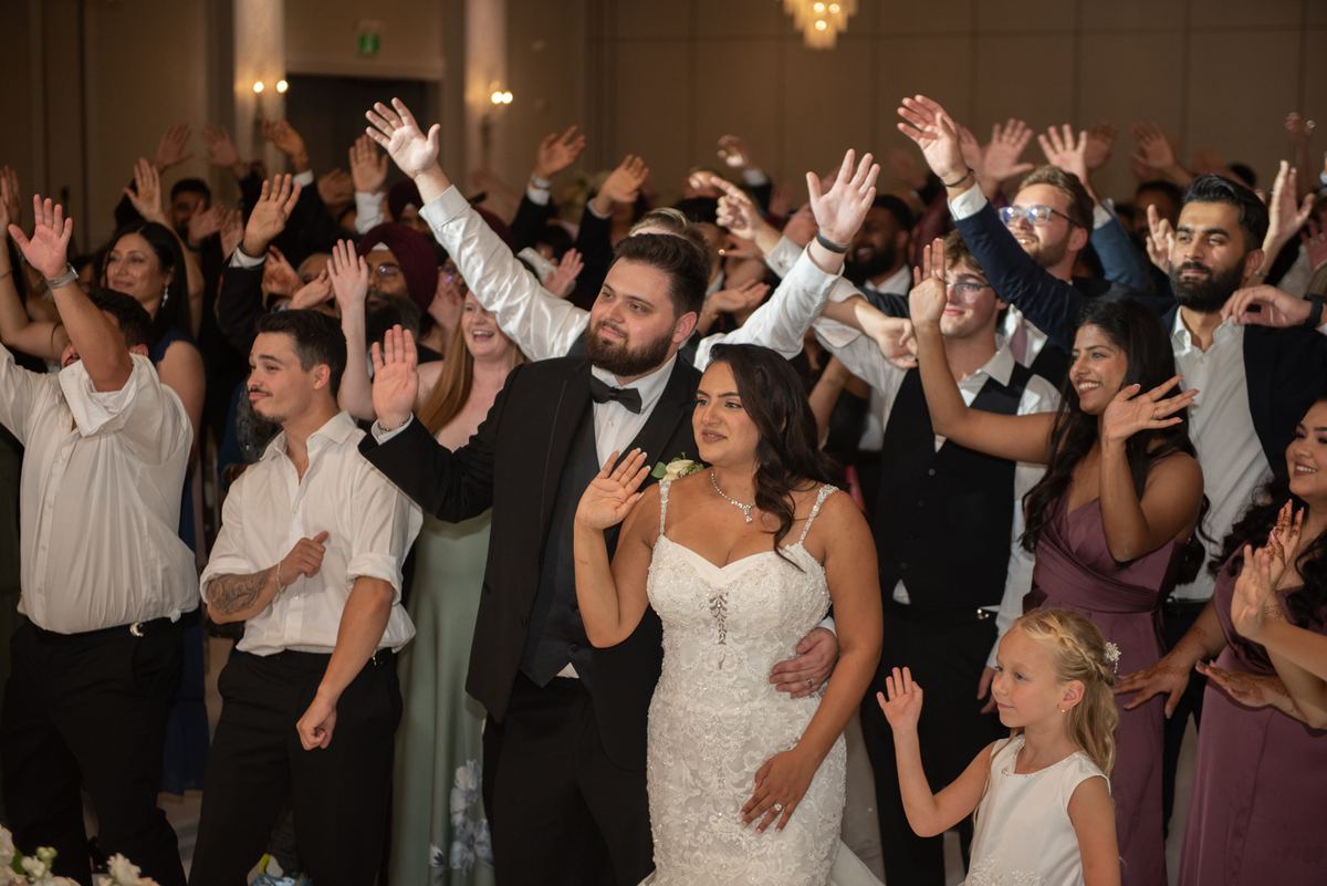 Bride and groom walk through cheering reception crowd, guests raising hands in celebration, highlighting festive atmosphere.