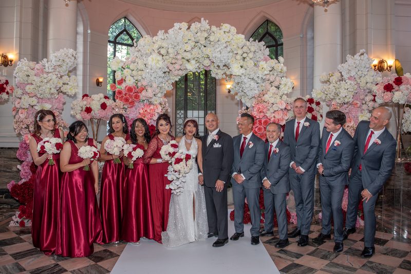 A bride in a white gown and groom stand under a floral arch with bridesmaids and groomsmen in formal attire.