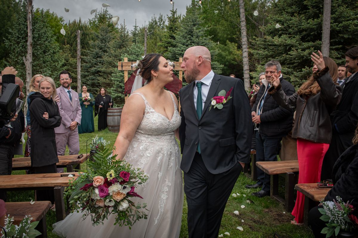 Bride and groom kiss at an outdoor wedding, guests cheer and toss petals in the air under a rustic wooden arch.