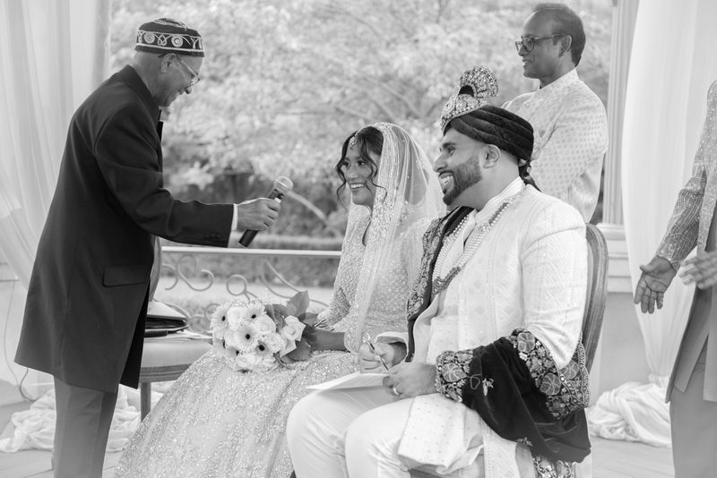 A bride and groom exchange vows outdoors under draped fabric and foliage, smiling as they sign documents.
