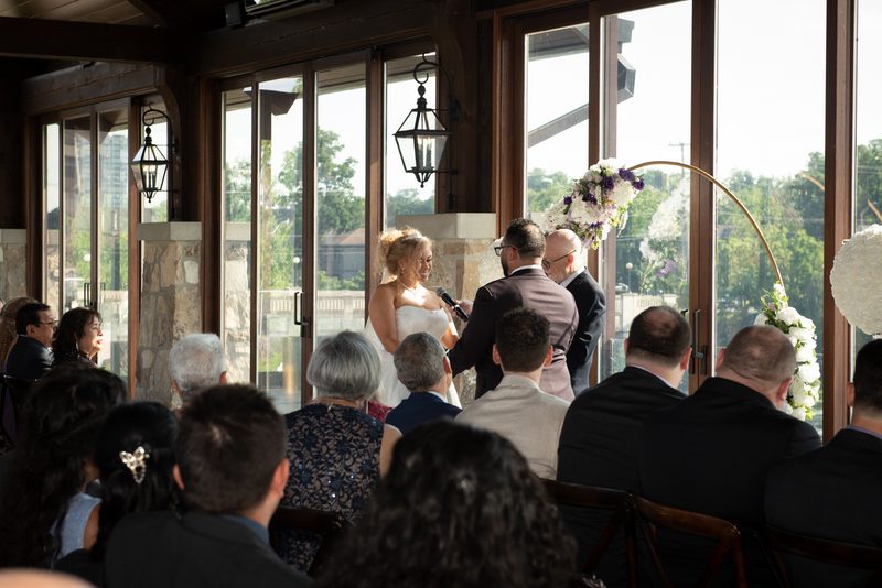 Bride and groom smile at altar as guests look on in natural light, surrounded by flowers and stone-and-wood decor.