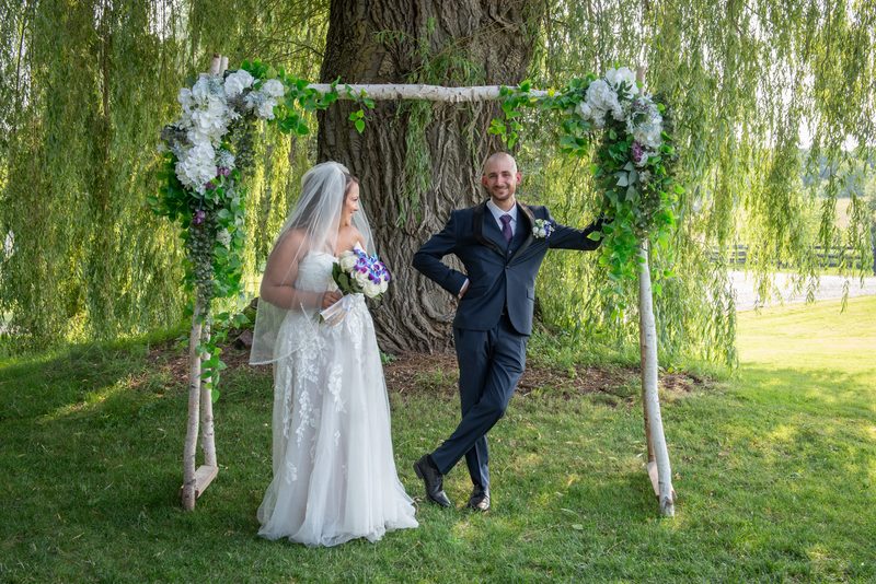 A bride and groom stand under a birch-branch arch adorned with hydrangeas, set against a willow tree.
