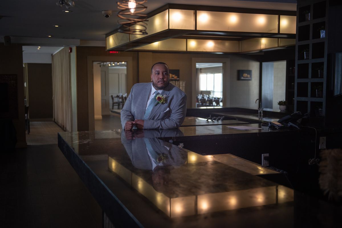 A groom in a light gray suit stands at a dark reception desk in a modern hotel lobby, his reflection visible.