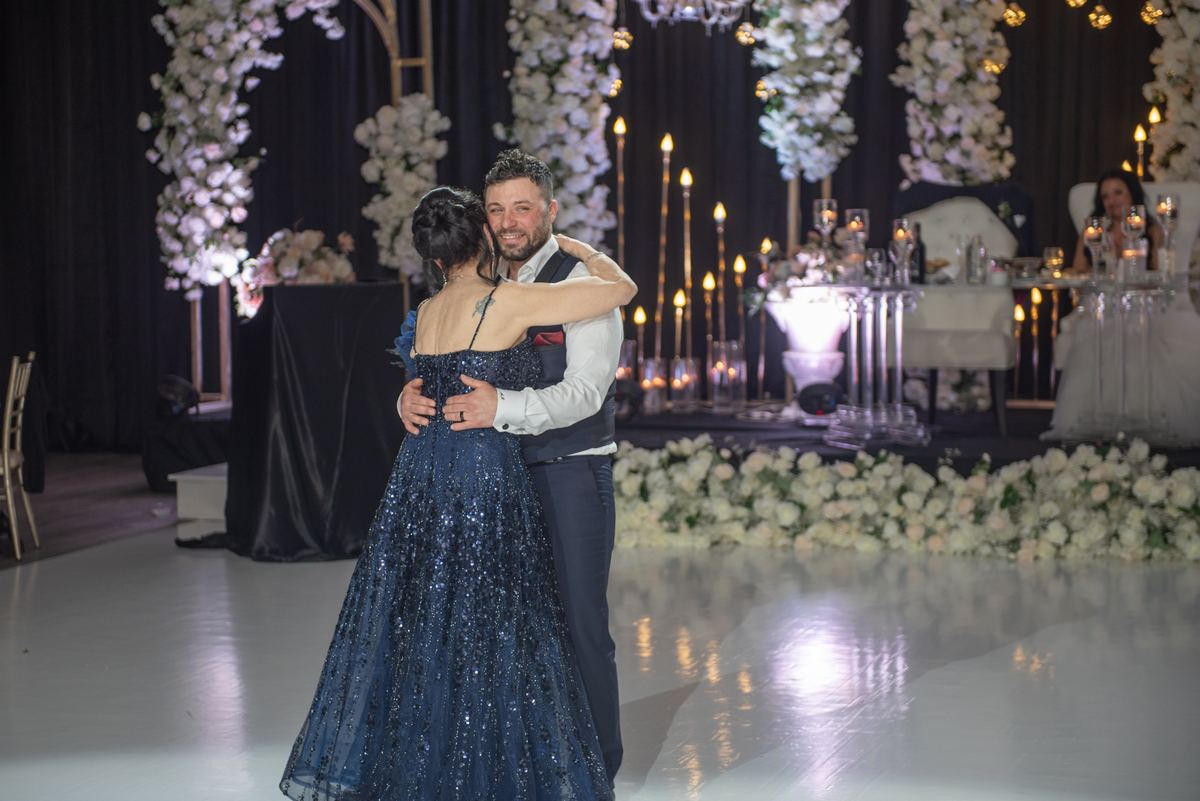 A couple dances on a glossy floor, surrounded by white flowers and candles, guests watching from tables.