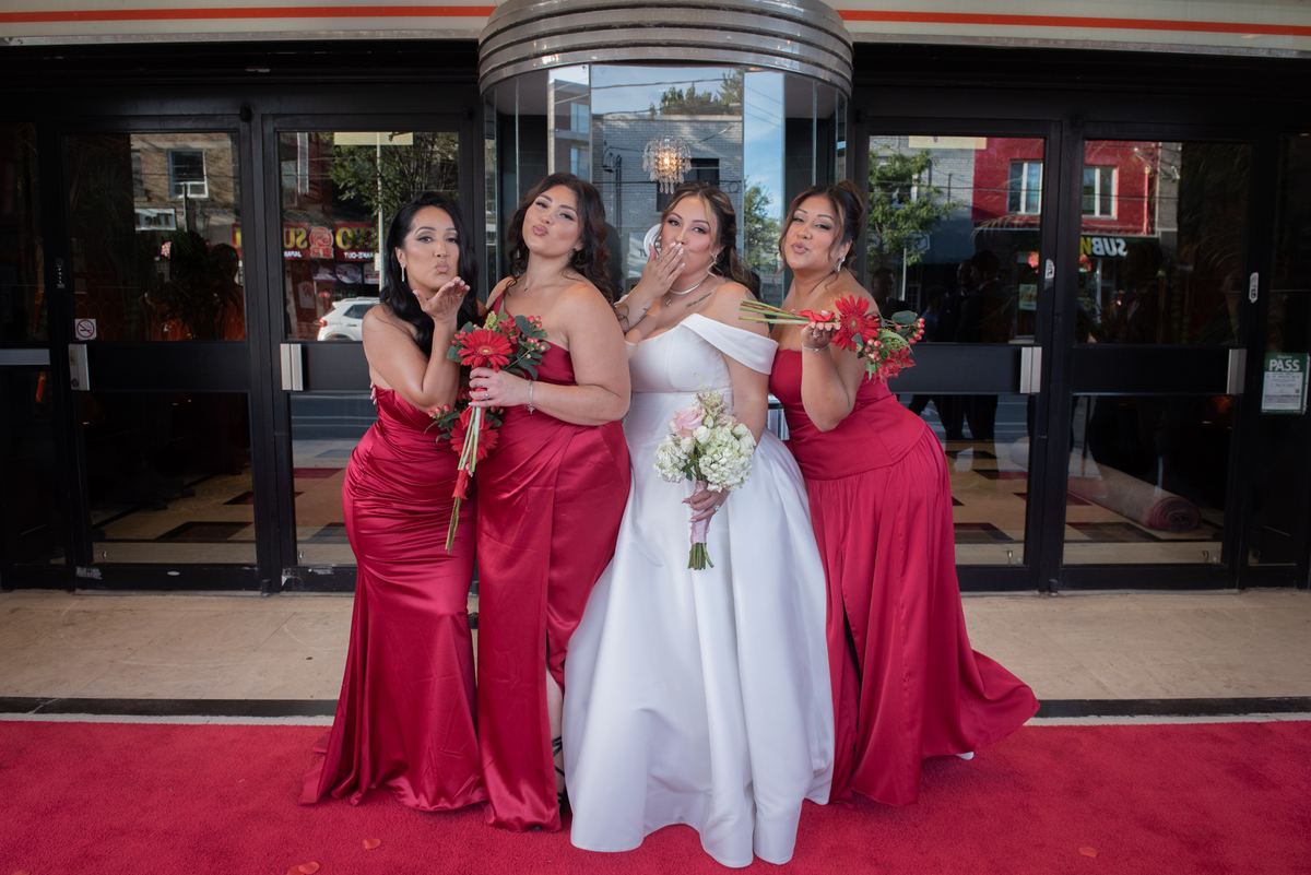 Four women, including a bride in white and bridesmaids in red, pose playfully with bouquets on a red carpet.