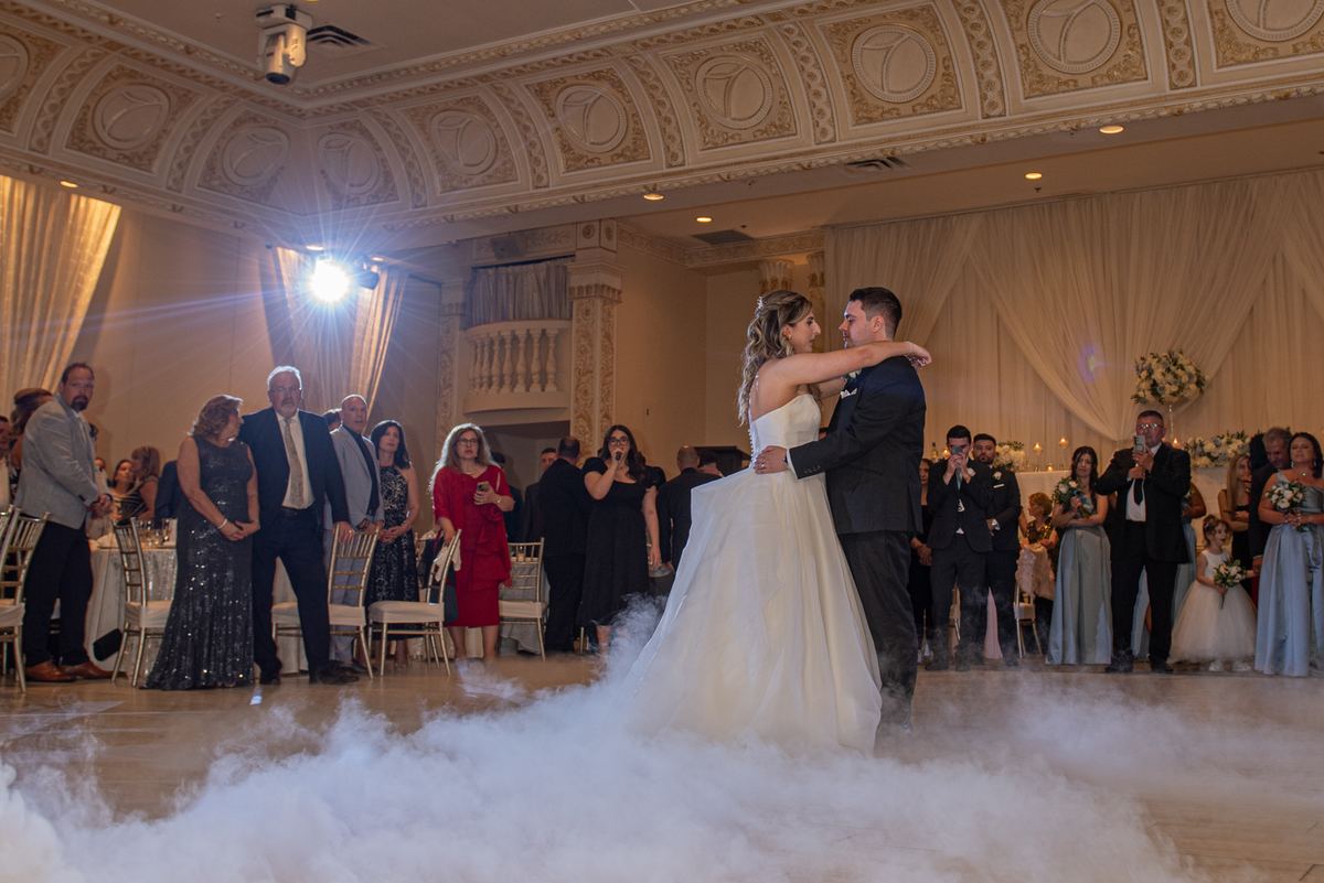 Bride and groom dance under spotlight in foggy ballroom with guests watching, surrounded by ornate drapes.