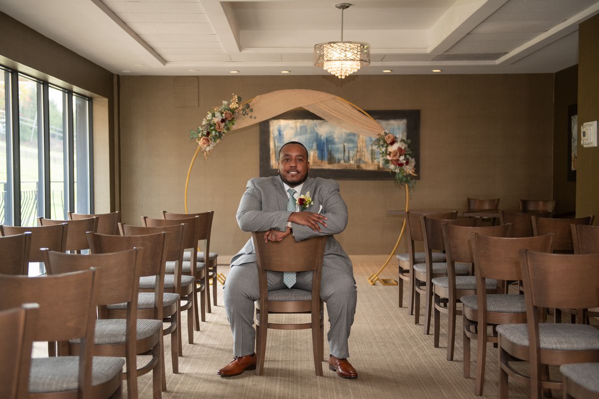 A groom in a gray suit sits confidently on a chair, smiling at the camera during a wedding ceremony.