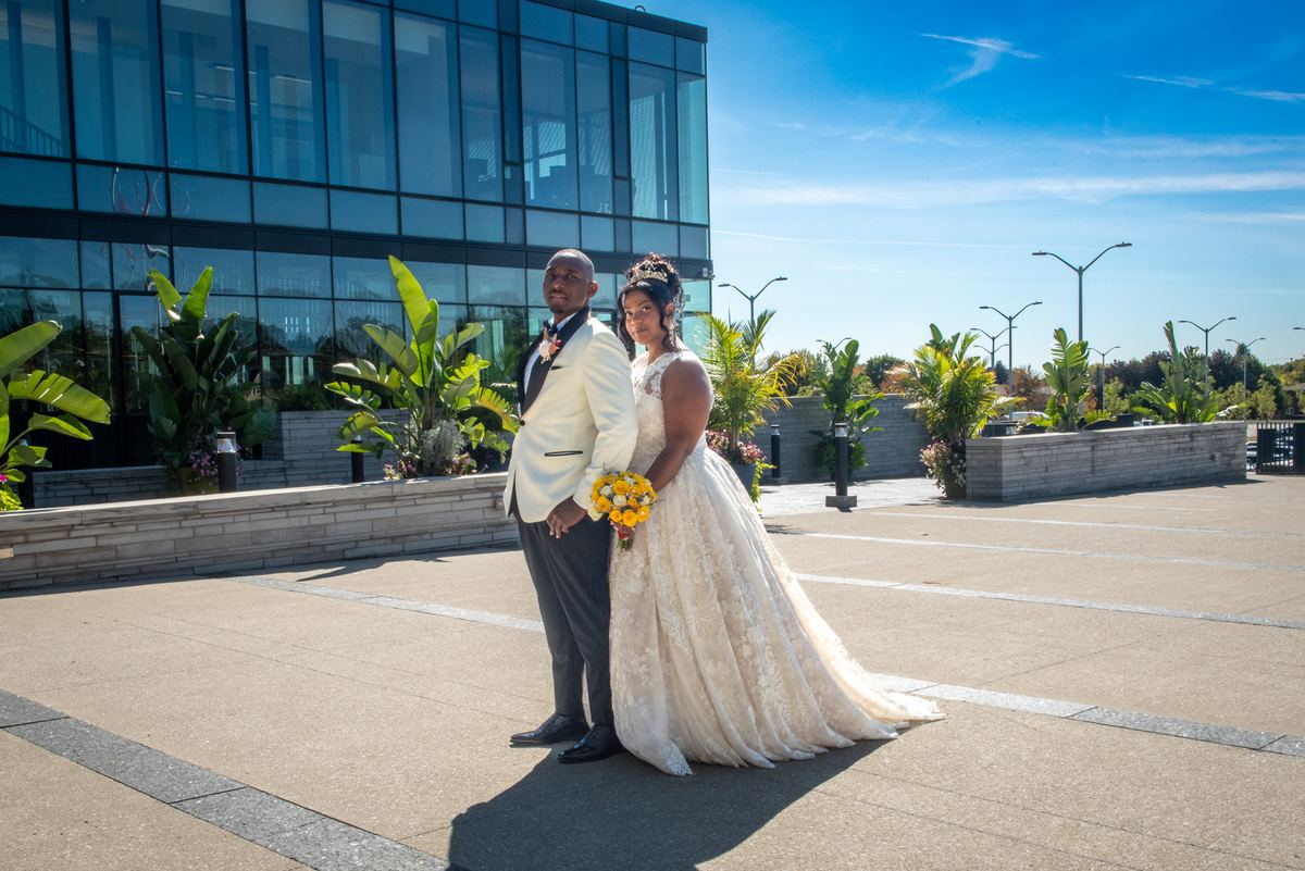 A bride in a lace gown and groom in a white tuxedo stand in front of a modern glass building under a clear blue sky.