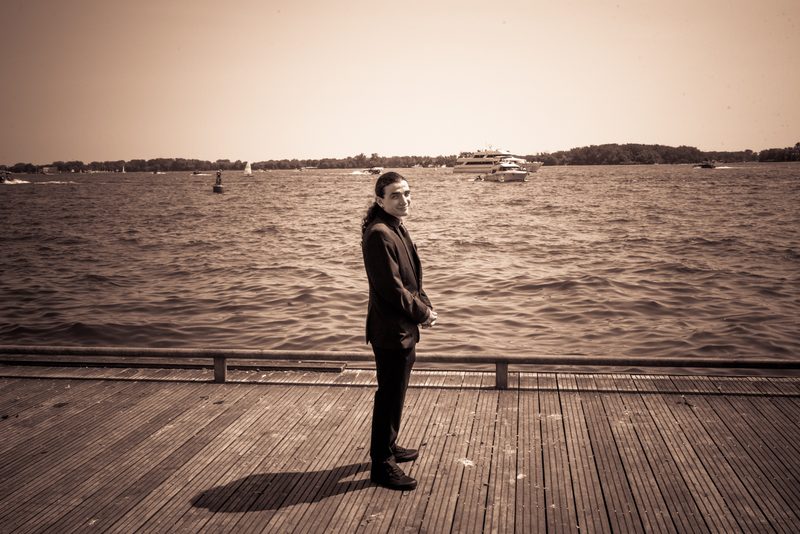 A man stands alone on a wooden pier, gazing out over a waterway filled with boats under a hazy, sepia-toned sky.