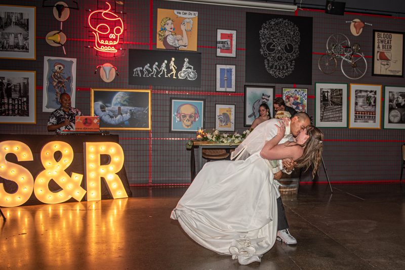 A bride and groom share an intimate kiss during their wedding dance, with warm marquee letters 'S&R' in the foreground.