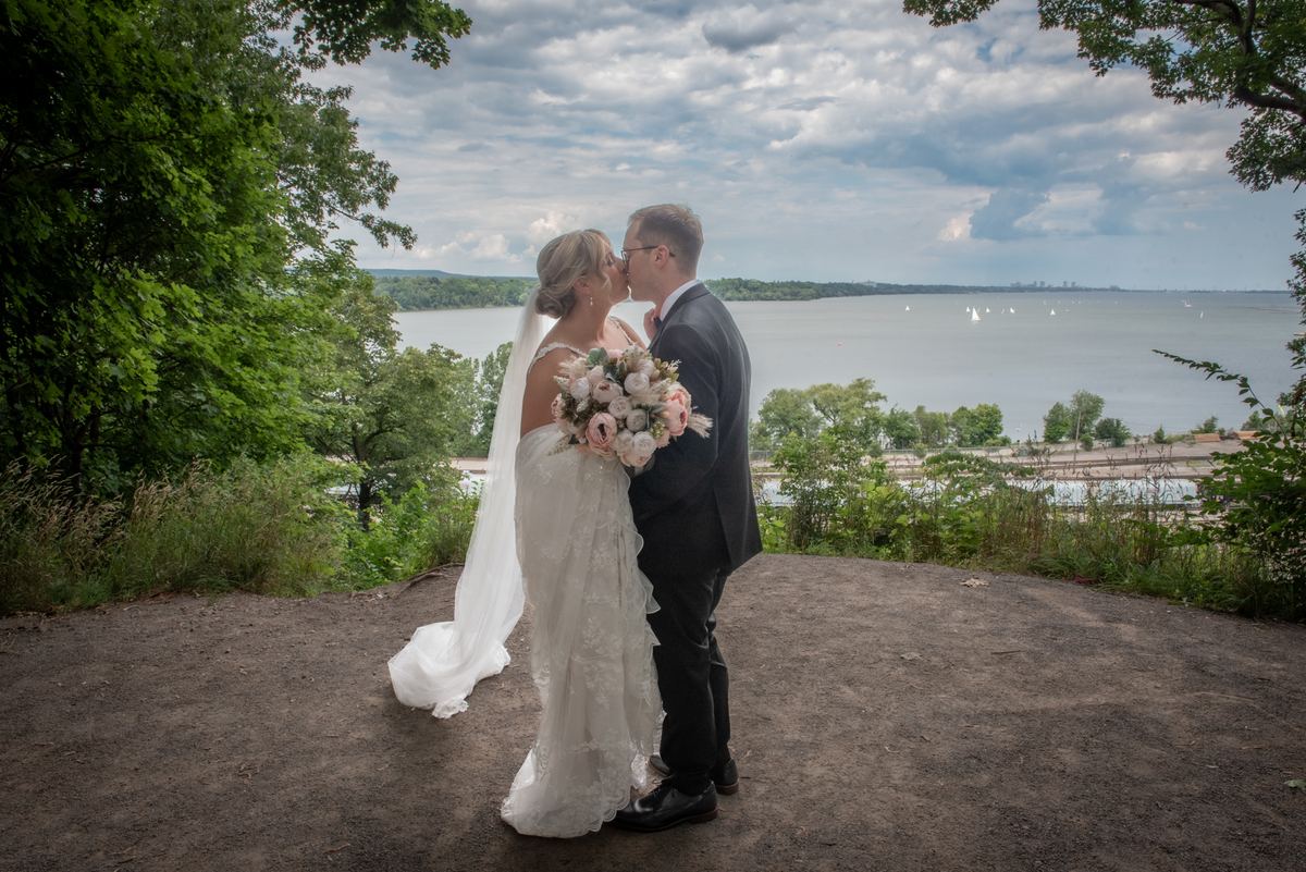 A bride and groom kiss at a dirt overlook, surrounded by greenery and a lake with sailboats, under a cloudy sky.