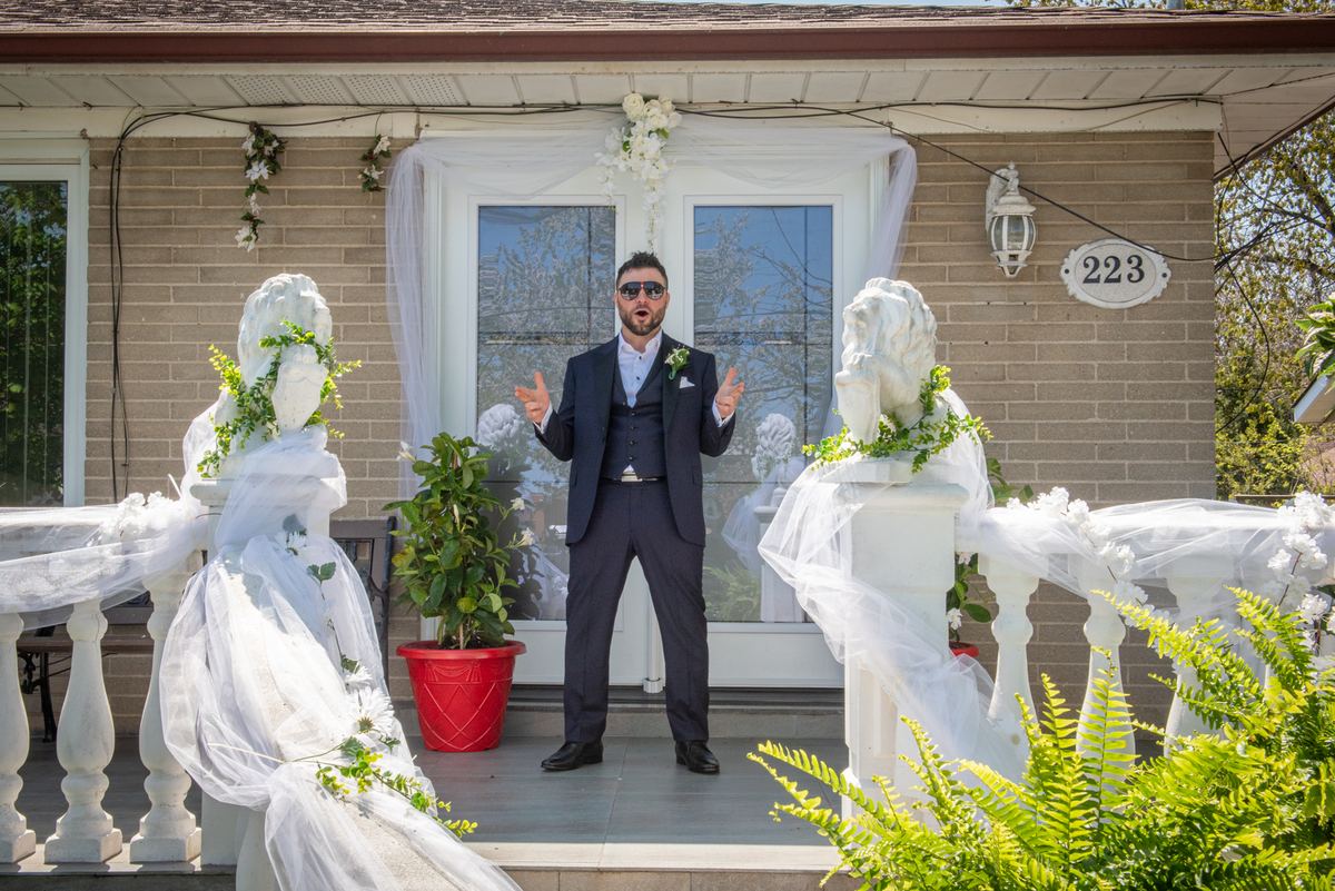 A man in a dark suit and sunglasses gestures animatedly on a wedding porch decorated with white veils and flowers.