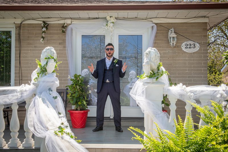 A man in a dark suit and sunglasses gestures animatedly on a wedding porch decorated with white veils and flowers.