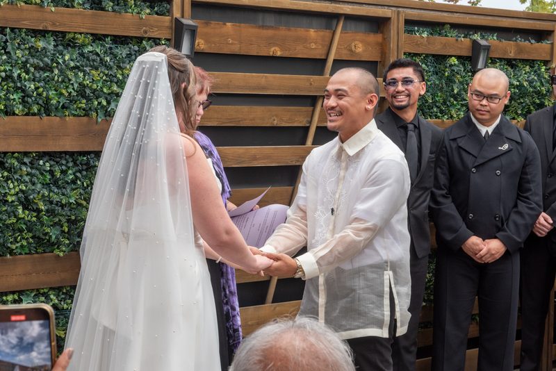 A bride and groom exchange vows outdoors, smiling as they hold hands, with the groom in traditional attire.
