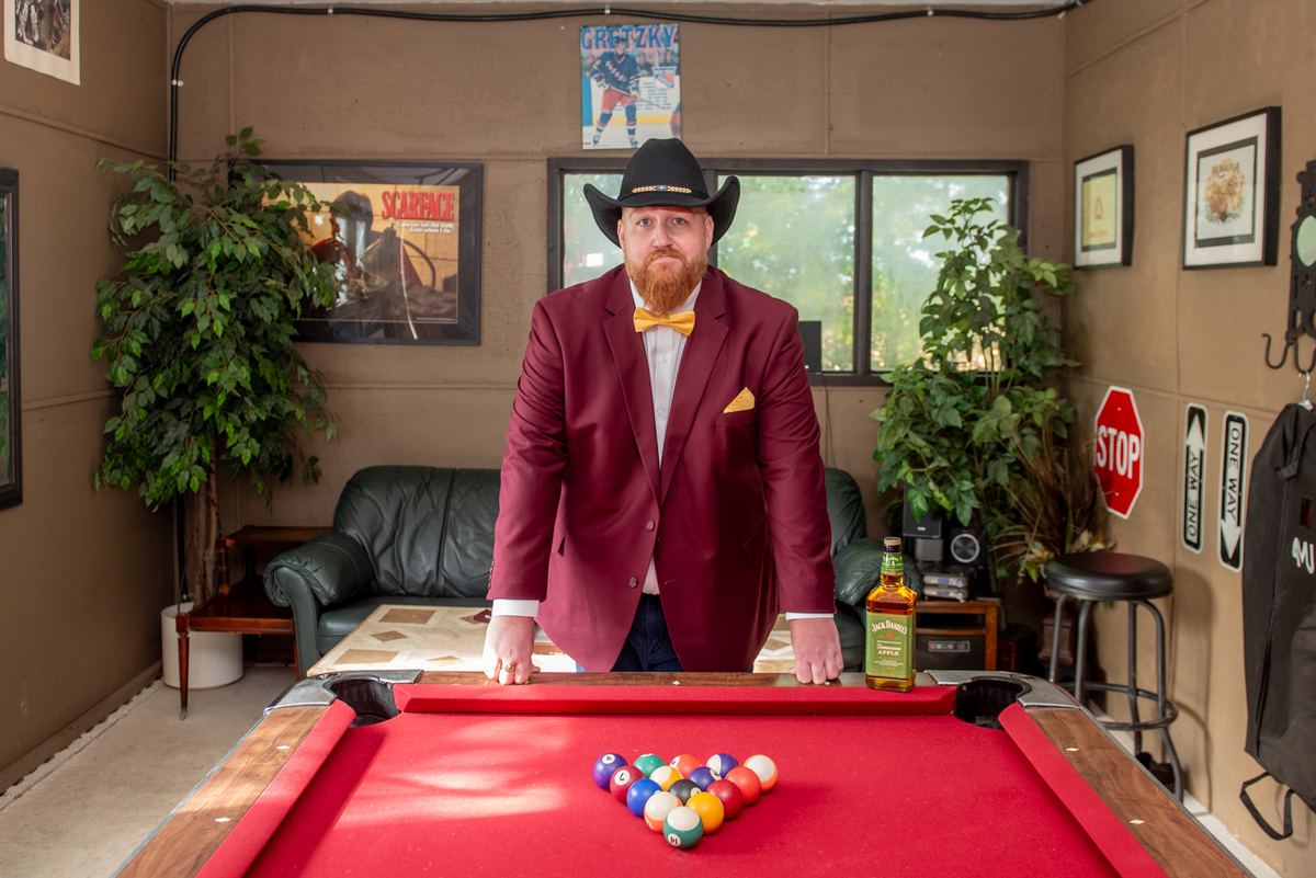 A man in a maroon suit and yellow bow tie stands behind a pool table with balls arranged in a triangle.