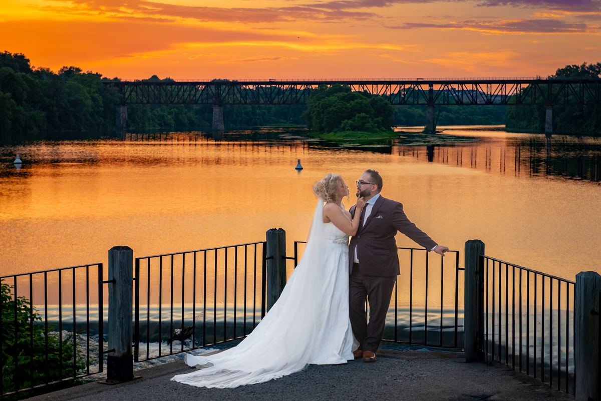 A bride touches her groom's face on a waterfront walk, their silhouettes against a fiery orange sunset mirrored in the river.