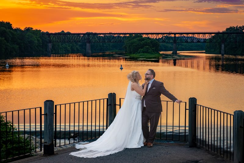 A bride touches her groom's face on a waterfront walk, their silhouettes against a fiery orange sunset mirrored in the river.