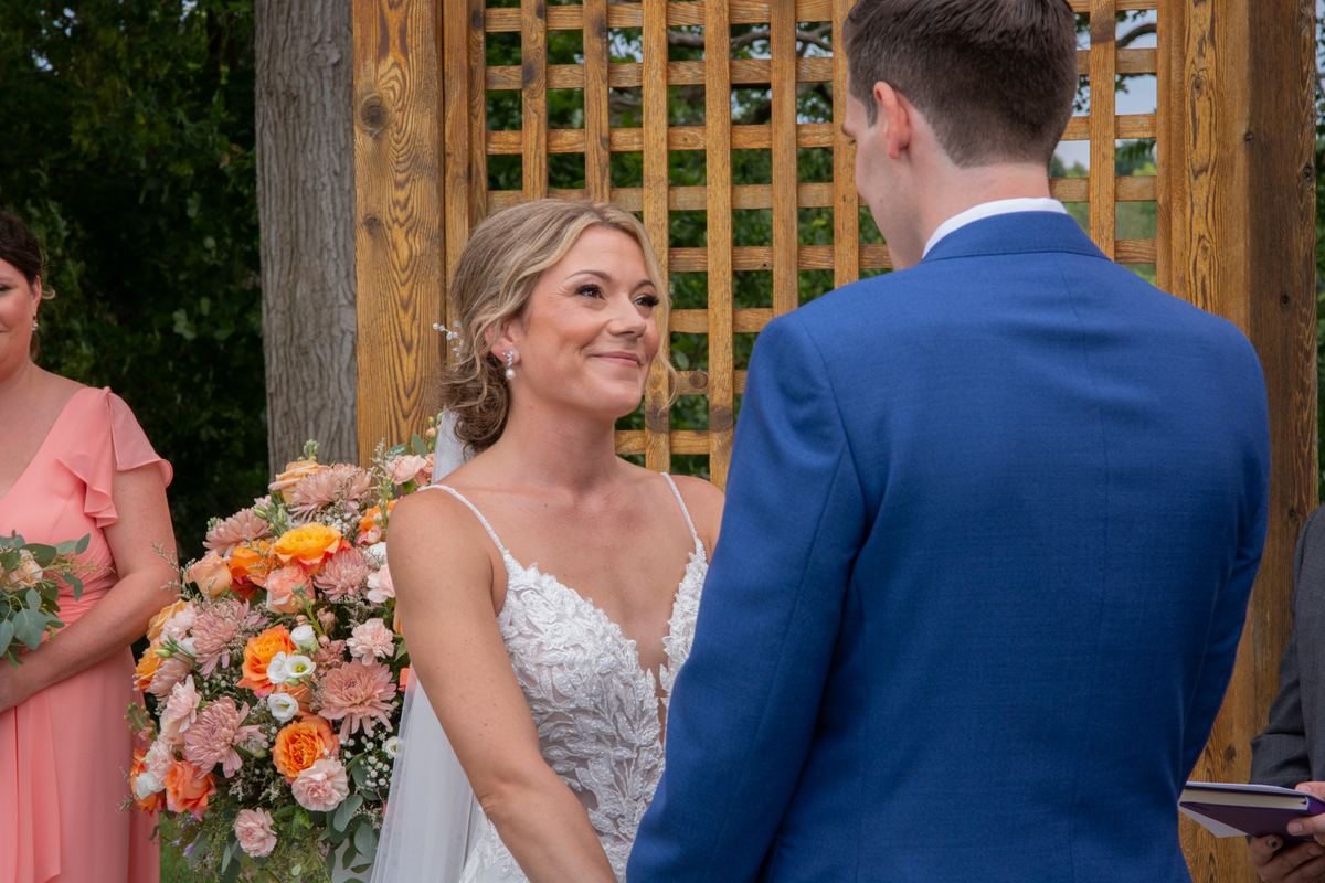 A bride in a lace gown holding her bouquet smiles at her groom in a blue suit at an outdoor ceremony before a wooden lattice.