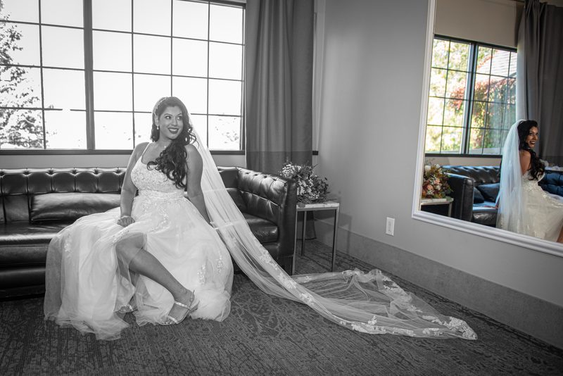 A bride sits on a leather sofa, smiling, with her veil trailing on the floor and her reflection showing in a mirror.