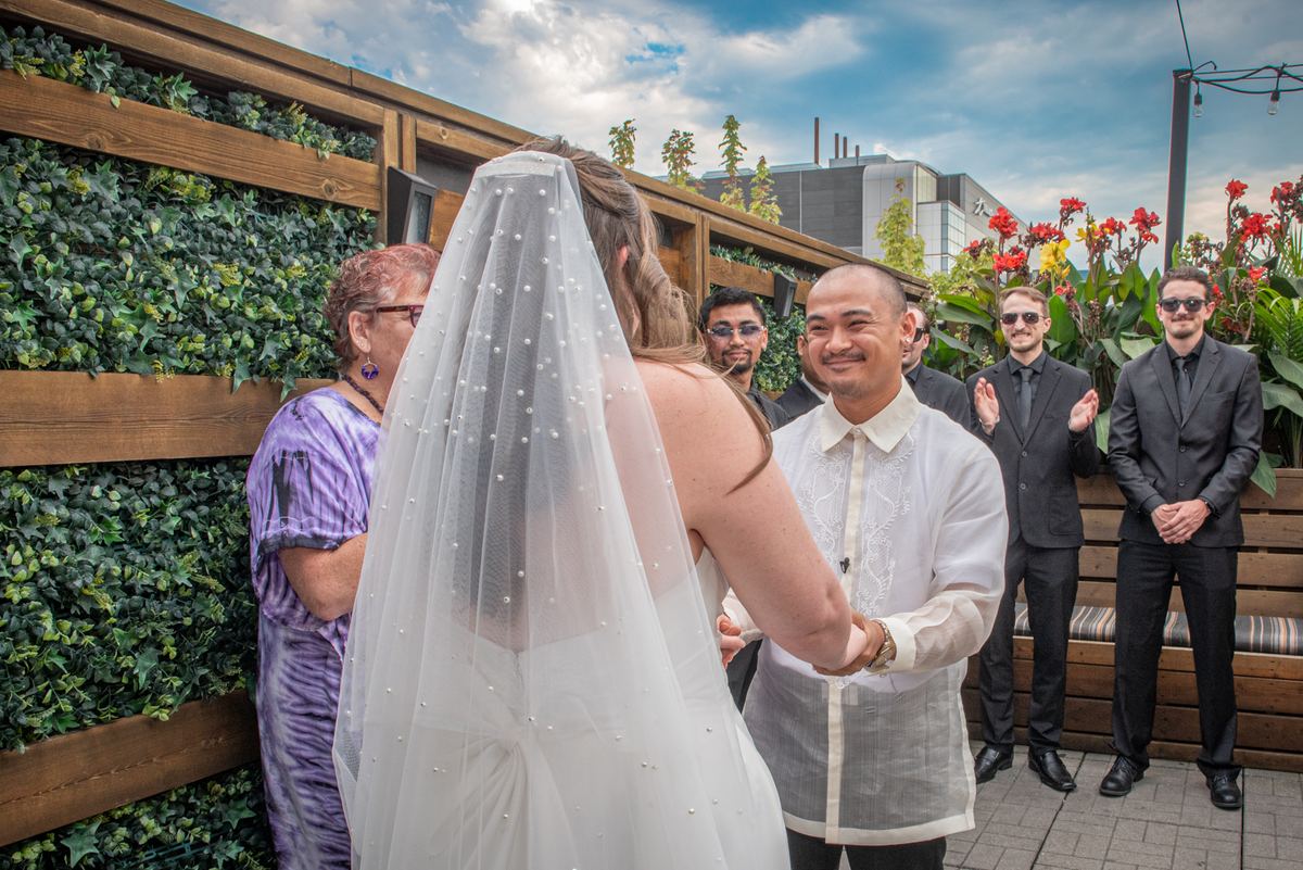 A bride and groom exchange vows outdoors, smiling as they hold hands in bright daylight with guests clapping.