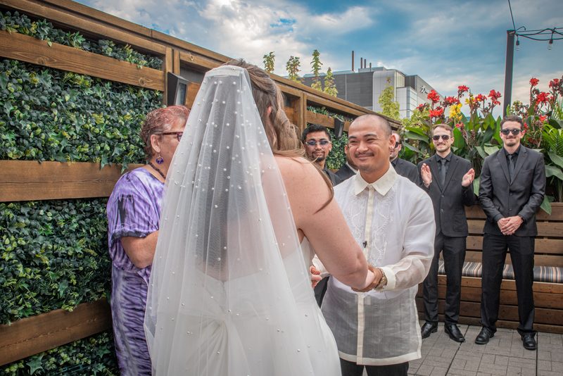 A bride and groom exchange vows outdoors, smiling as they hold hands in bright daylight with guests clapping.