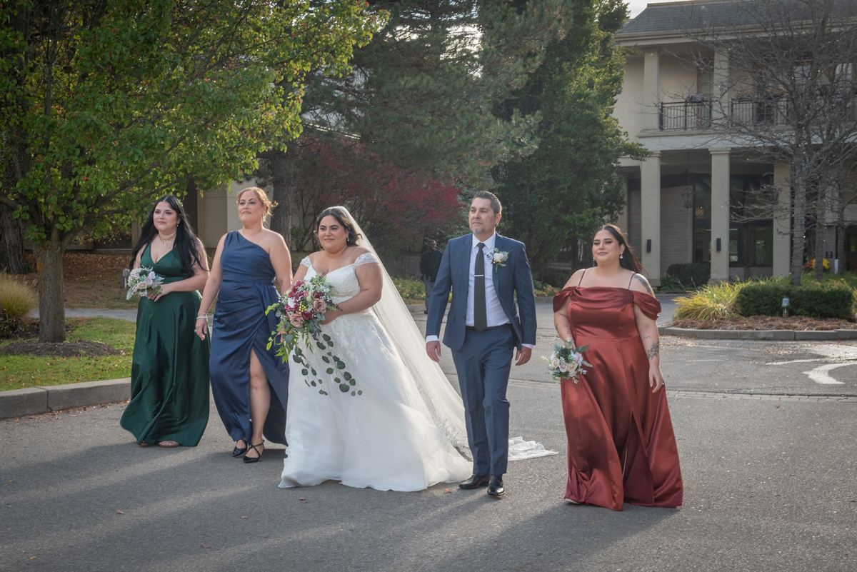 A bride in white walks with her groom and wedding party along a tree-lined path bathed in golden afternoon light.