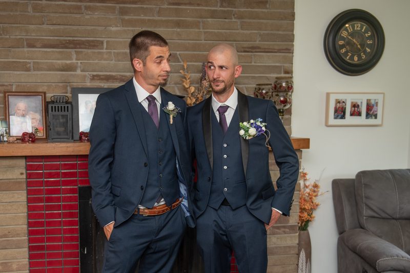 Two men in navy suits stand by a brick fireplace, sharing an amused glance with boutonnieres on their lapels.