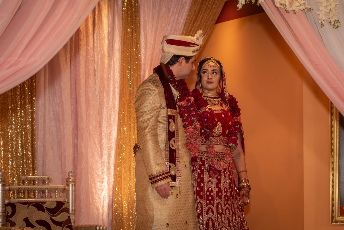 A newlywed couple in traditional attire stands together, gazing affectionately, framed by festive drapery.
