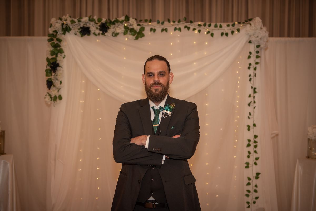 A groom in a dark suit with green accents stands confidently before a wedding arch decorated with white drapery.