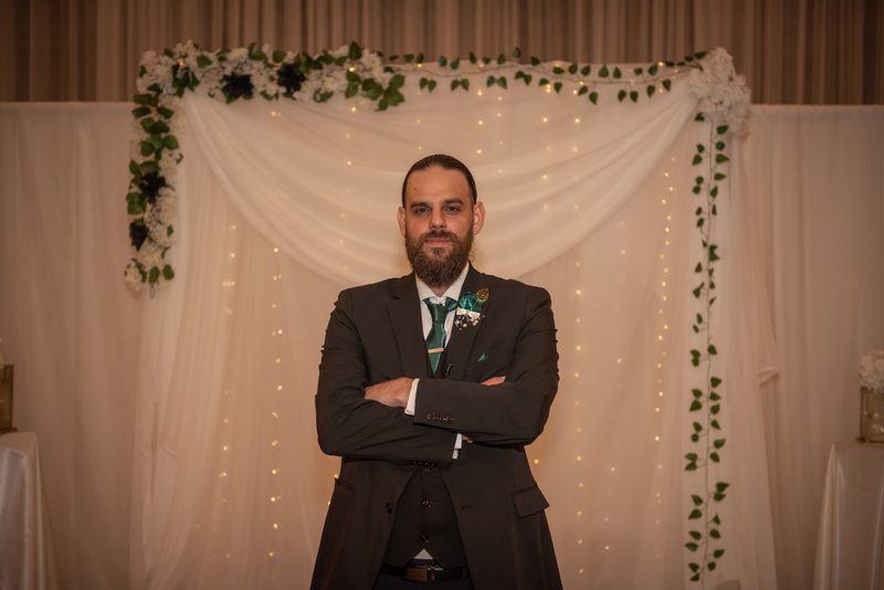A groom in a dark suit with green accents stands confidently before a wedding arch decorated with white drapery.