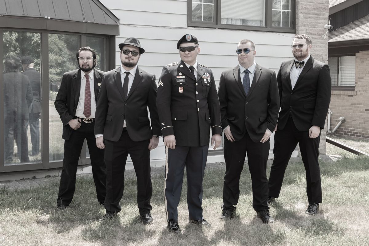 Five formally dressed men, including one in military uniform, pose outdoors in front of a building under bright sunlight.