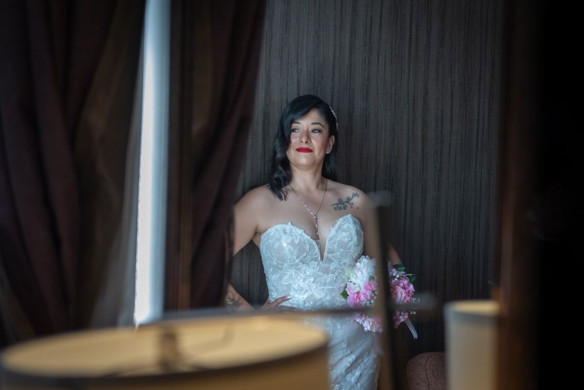 A bride in a lace-strapped gown holds flowers, her reflection visible in a mirror behind objects like lampshades.