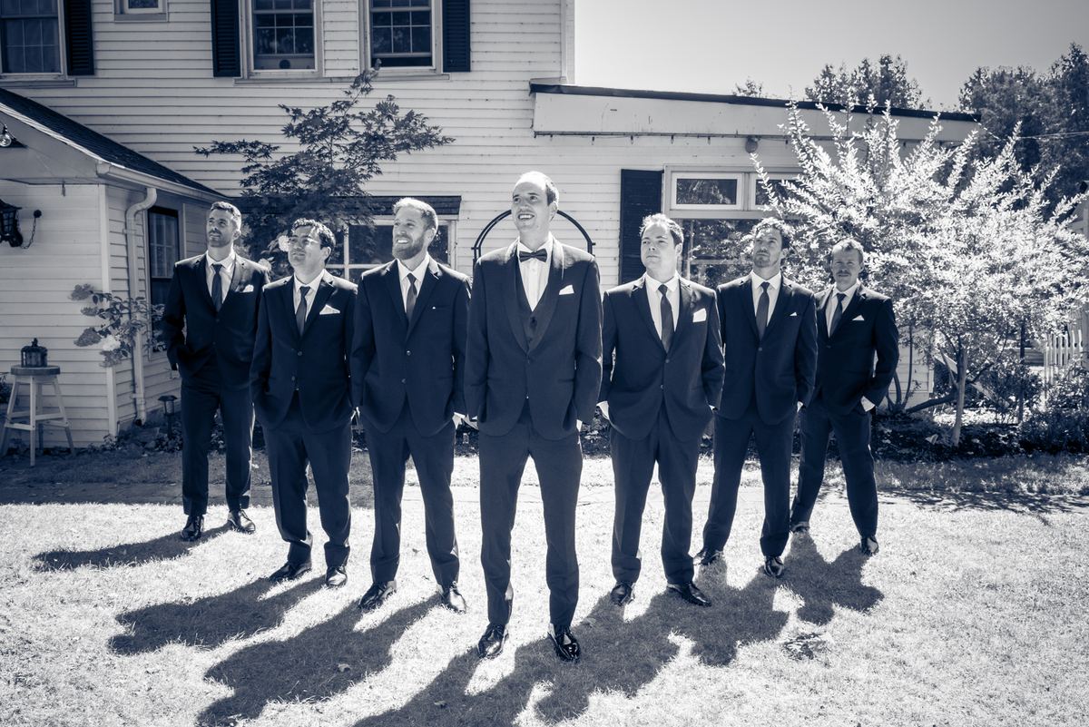 A groom and his groomsmen stand in formal suits on a sunlit lawn in front of a house, shadows stretching long behind them.