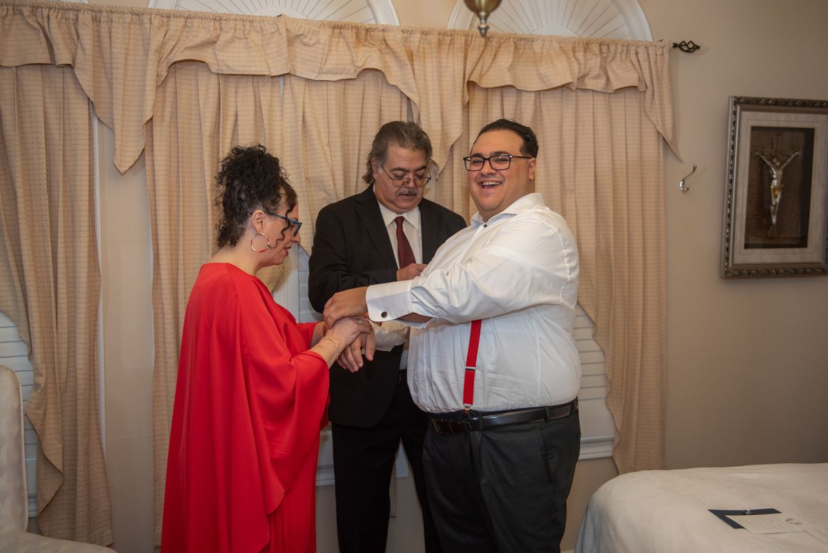 A happy couple in formal attire stands together as a man smiles nearby in a room with religious decor.
