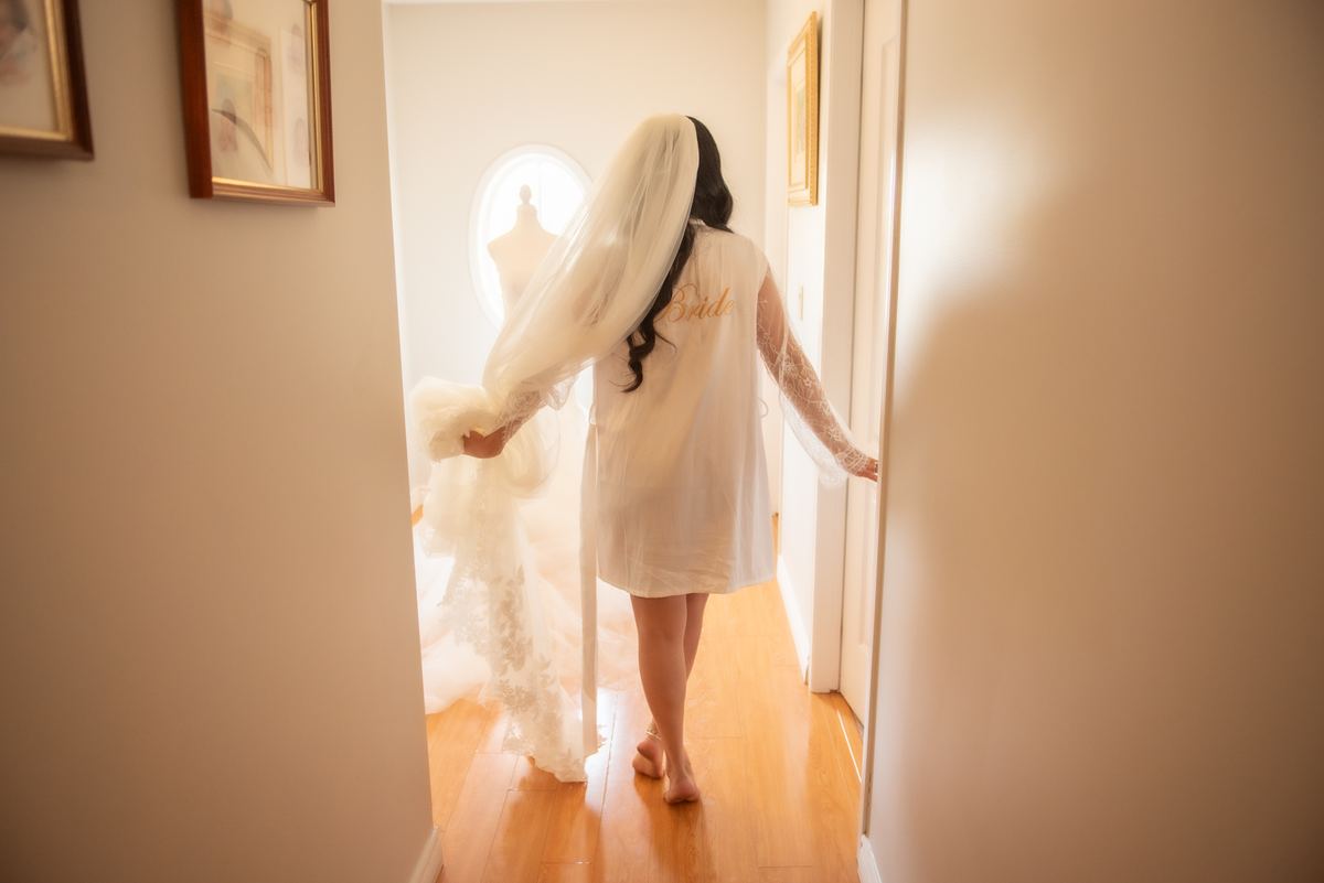 A bride in a white robe walks barefoot toward a bright doorway with her wedding gown displayed inside.