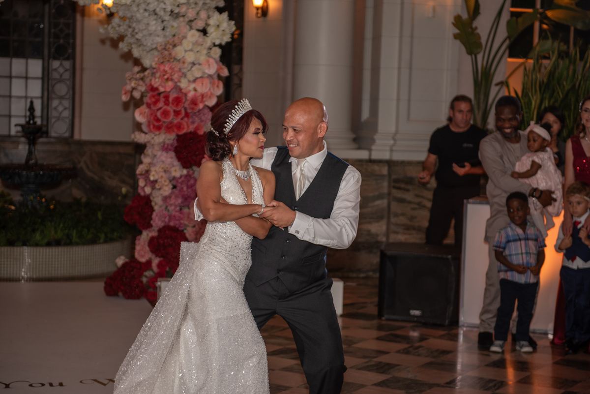 A bride and groom dance joyfully in a grand ballroom, surrounded by smiling guests and family members.