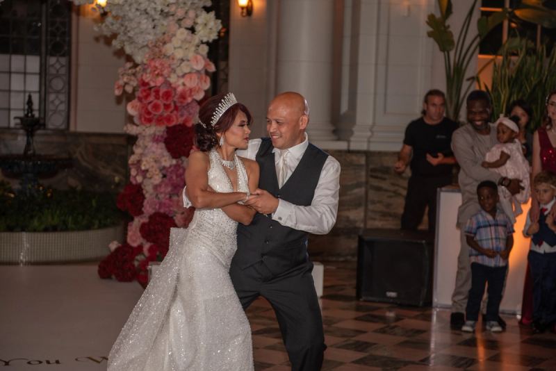 A bride and groom dance joyfully in a grand ballroom, surrounded by smiling guests and family members.