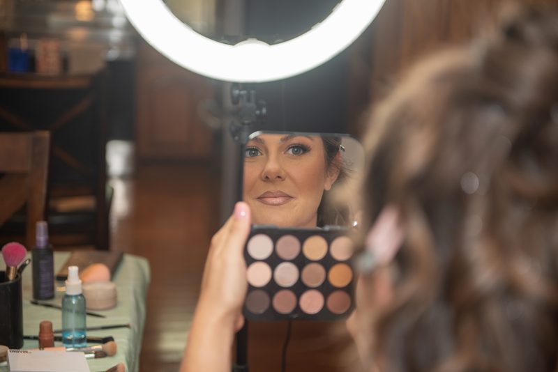 A woman applies makeup in front of a mirror, illuminated by a bright ring light, focusing on her beauty routine.