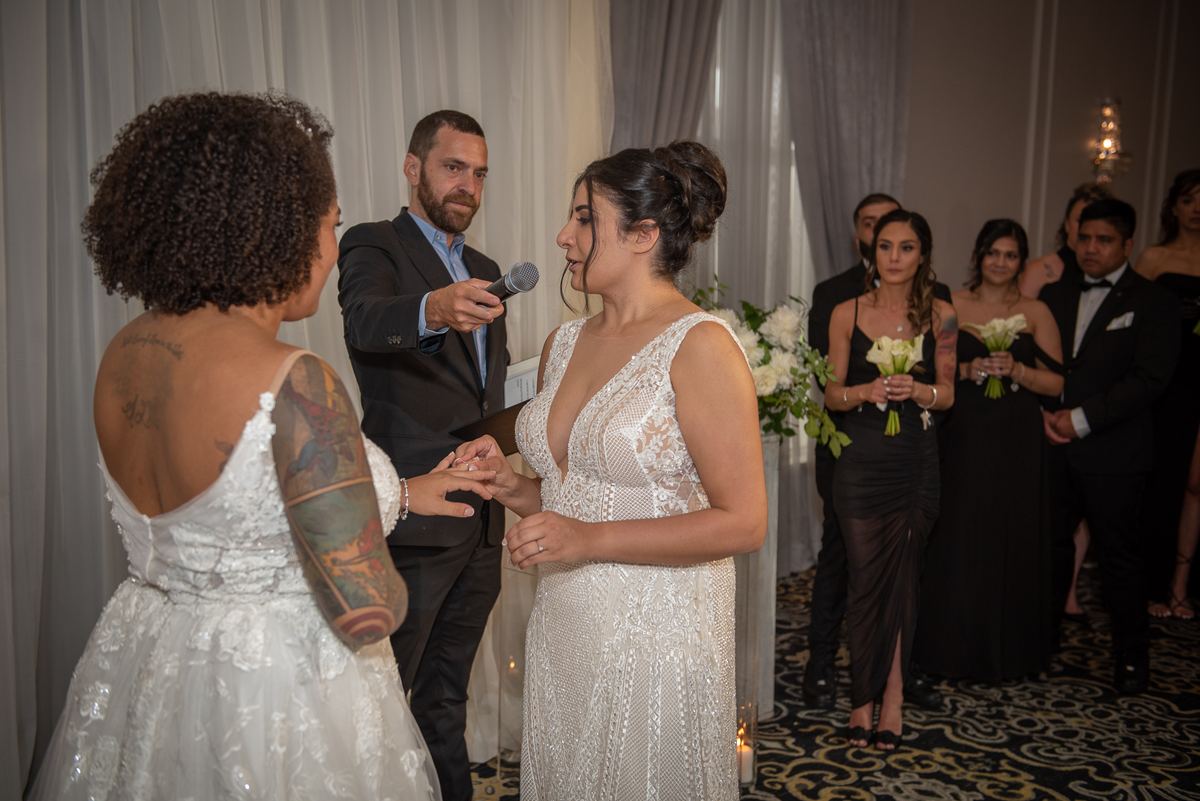 Two brides exchange rings as an officiant speaks, surrounded by formally dressed guests in a warmly lit setting.