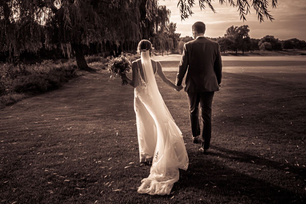 A bride and groom walk hand-in-hand across a grassy field at sunset, their figures silhouetted against the sky.