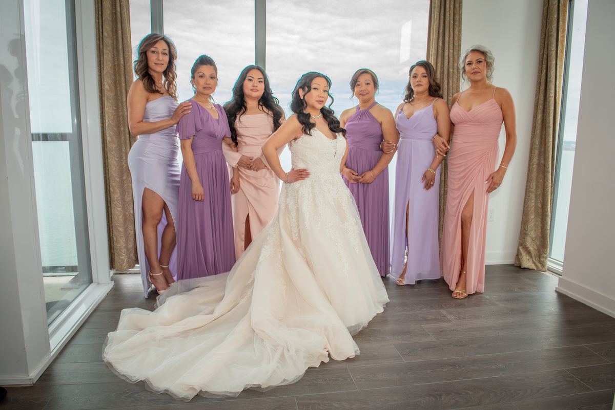 A bride in white stands confidently with six bridesmaids in pastel dresses near large windows, illuminated by soft light.