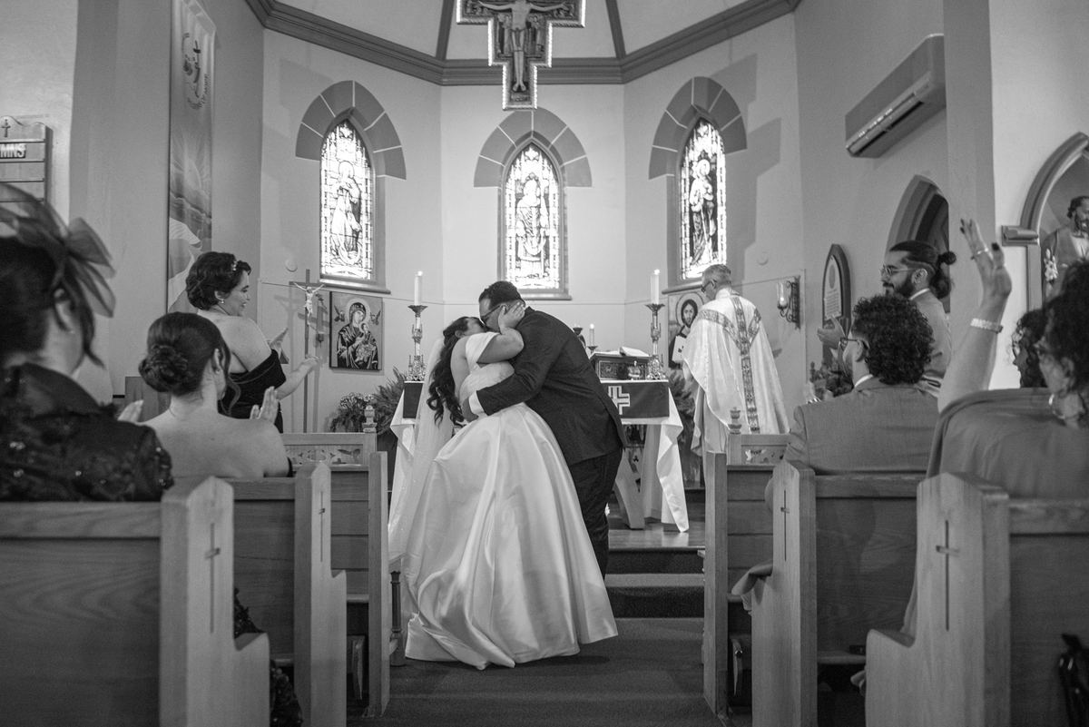 A bride and groom embrace at an altar surrounded by guests and clergy, illuminated by stained-glass light.