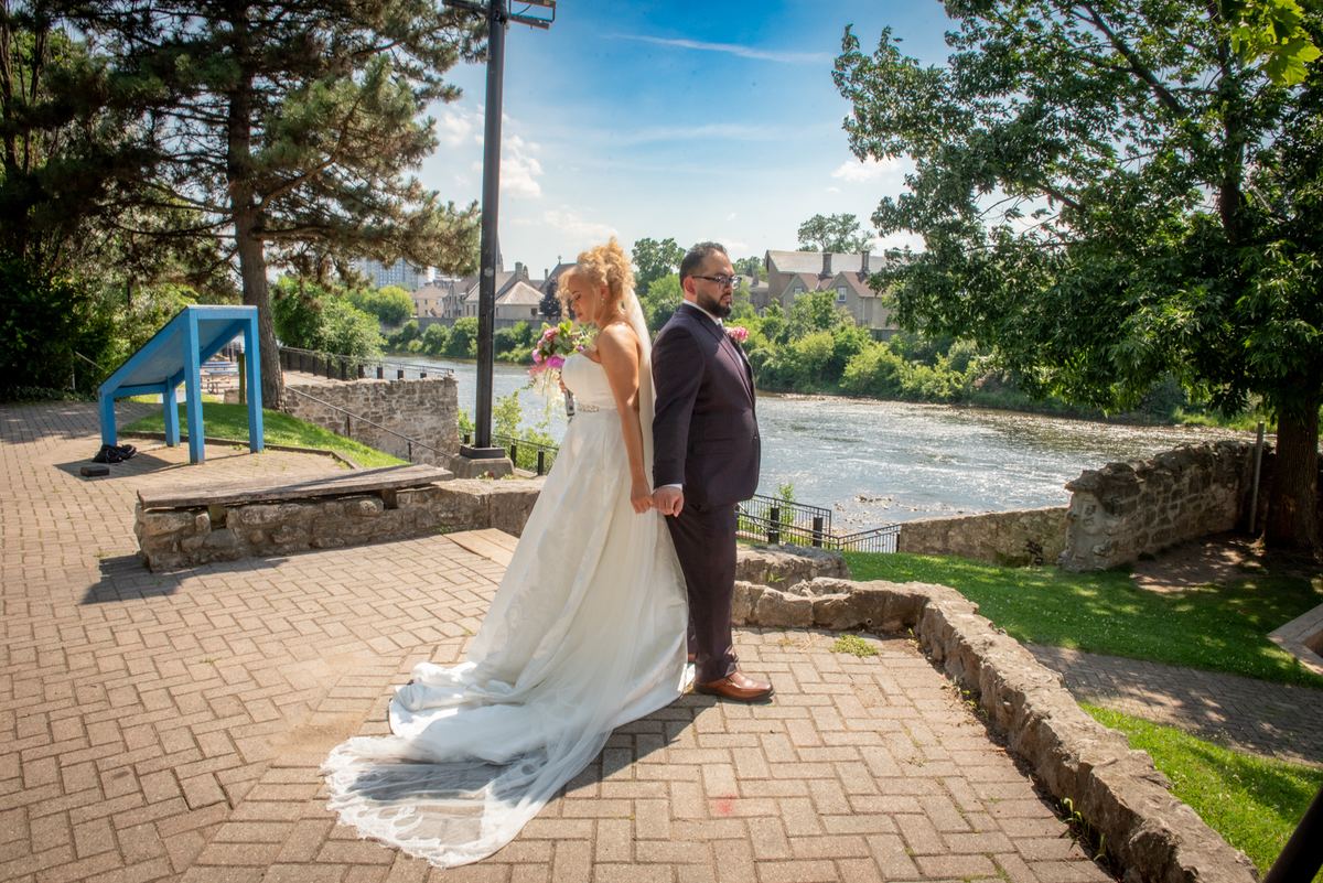 Bride and groom stand on stone overlook by river, hands clasped, under sunny sky with dappled light from trees.