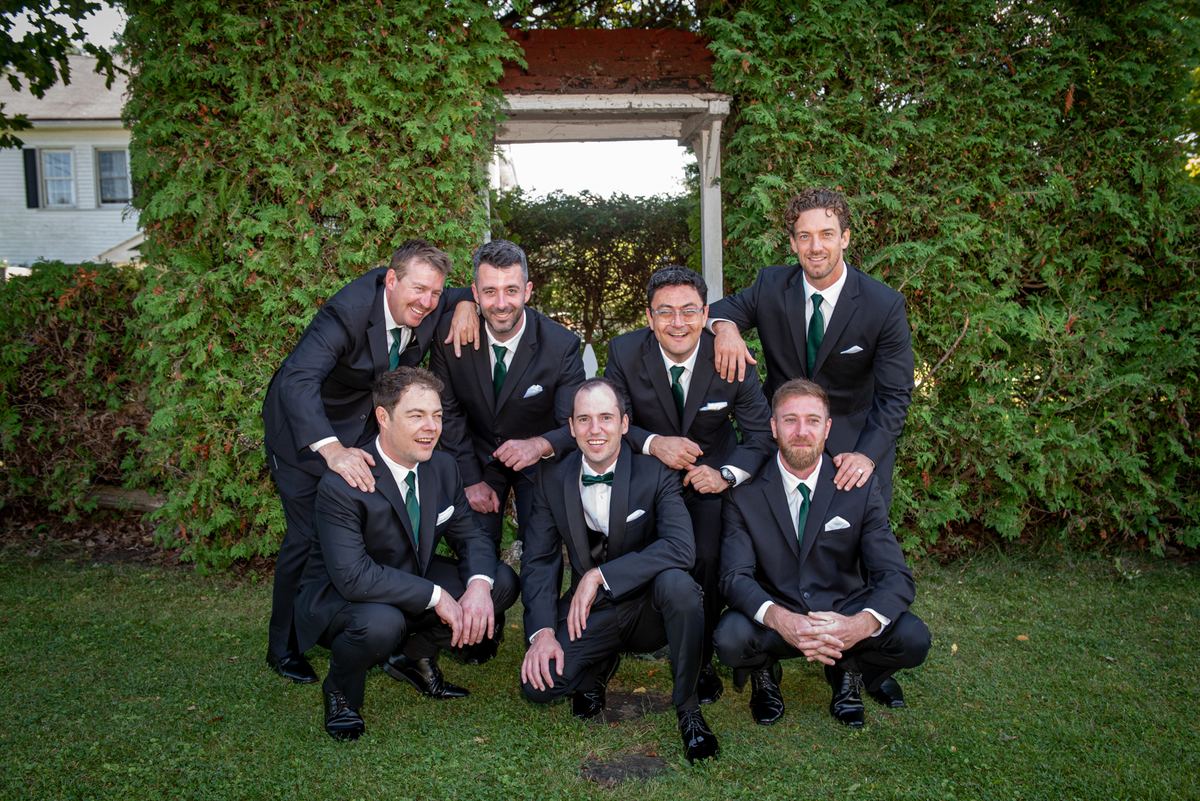 Seven groomsmen in black suits and green ties pose joyfully on a grassy lawn under trees, embracing.