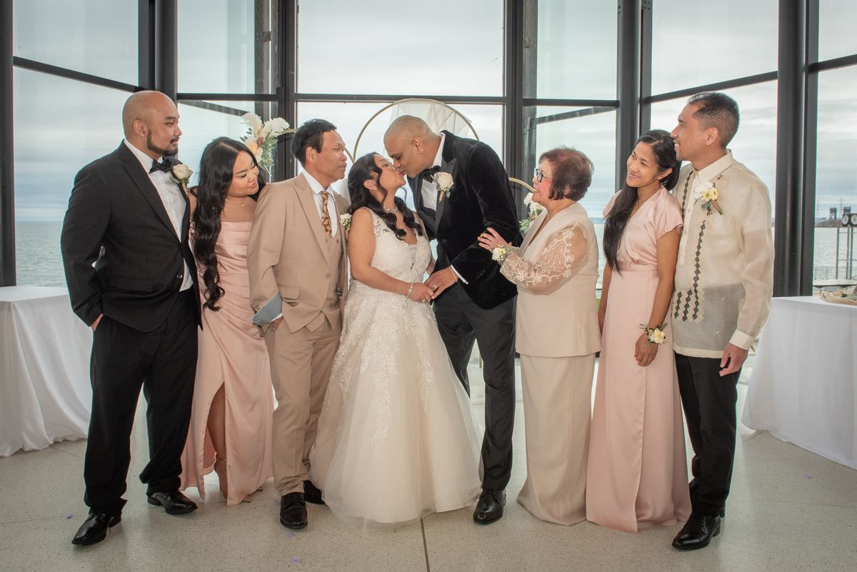 Bride and groom kiss surrounded by family in modern space with sea view, illuminated by natural light.